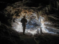 Bildet viser grottepassasje med dryppstein (stalaktitter i taket og stalagmitter på golvet) og en person med grotteutforskningsutstyr. Okshola-Kristihola, Fauske, Nordland.
