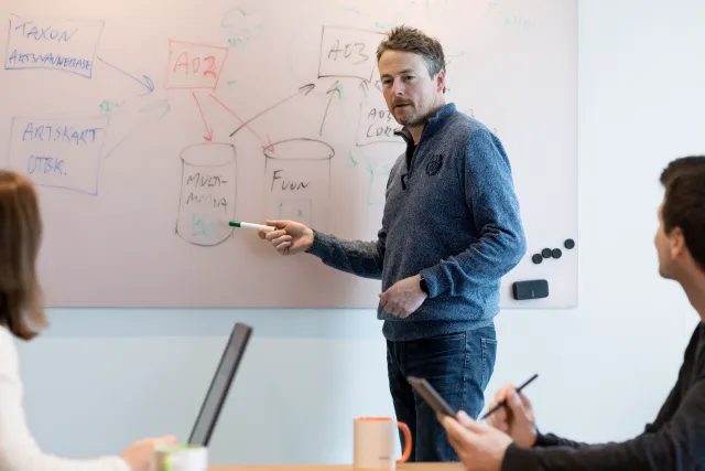 Ansatt forklarer og peker på tavle under en faglig gjennomgang, med kolleger til stede.