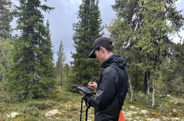 Mann i turklær står med ipad i skog for å kartlegge.