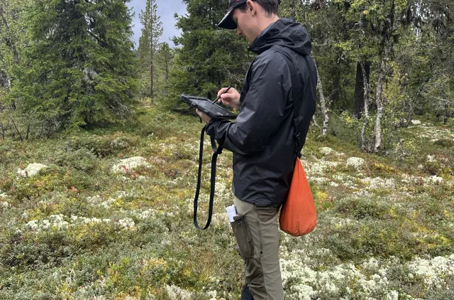 Mann i turklær står med ipad i skog for å kartlegge.