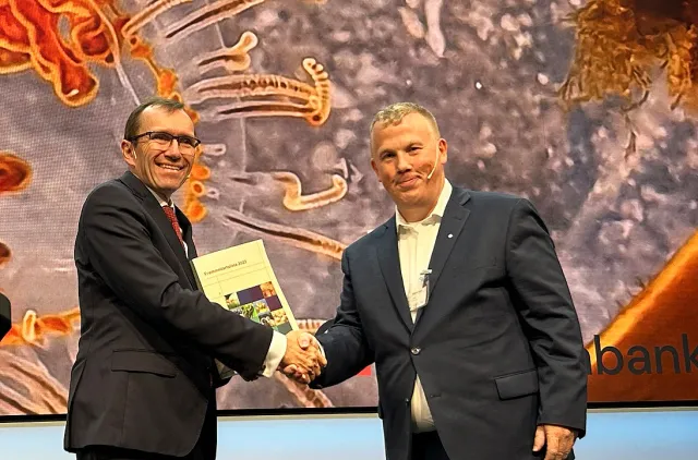 To menn i dress på en scene og det overrekkes en bok.